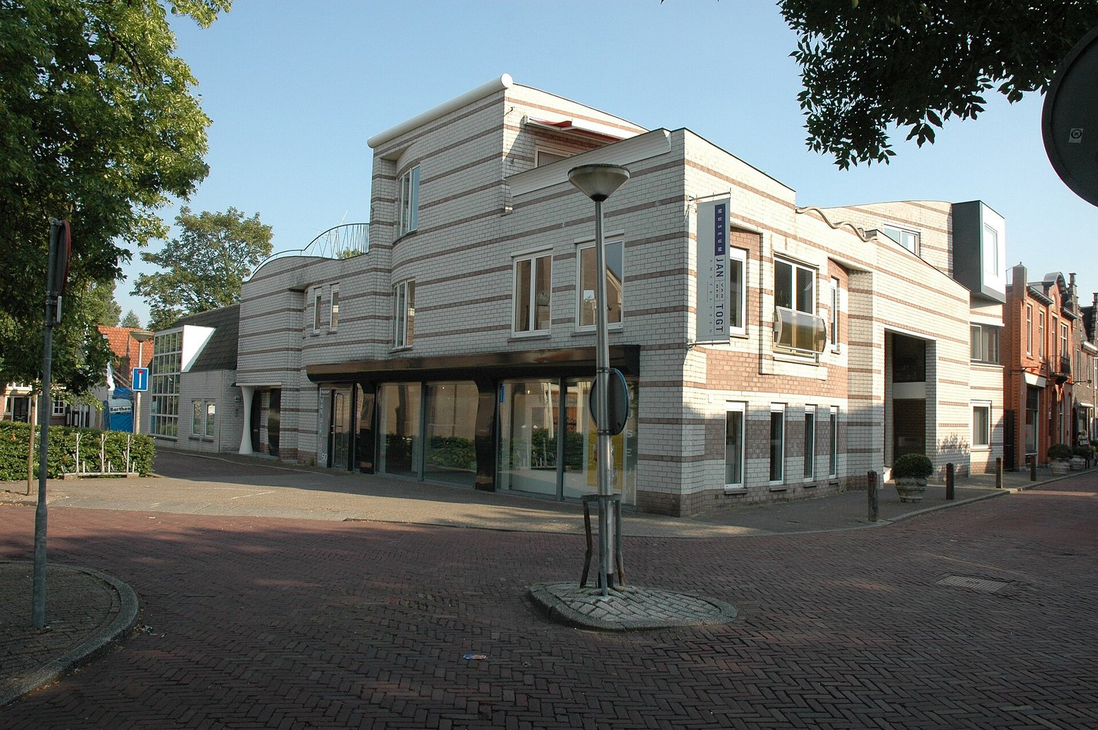 Hell gestreiftes Backstein-Museumsgebäude an einer gepflasterten Straßenecke, mit großen Schaufenstern im Erdgeschoss und einem senkrechten Namensschild an der Fassade.