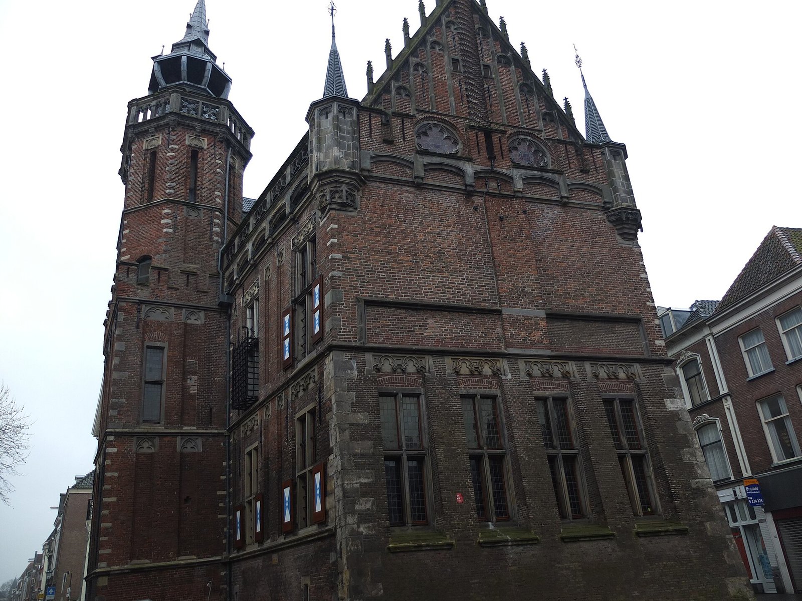 Laatgotisch bakstenen raadhuis met links een hoge toren met open lantaarn, een puntgevel met pinakels en torentjes, en onderaan grote vensters met glas in lood en rood-wit-blauwe luiken, gezien vanaf de straat.