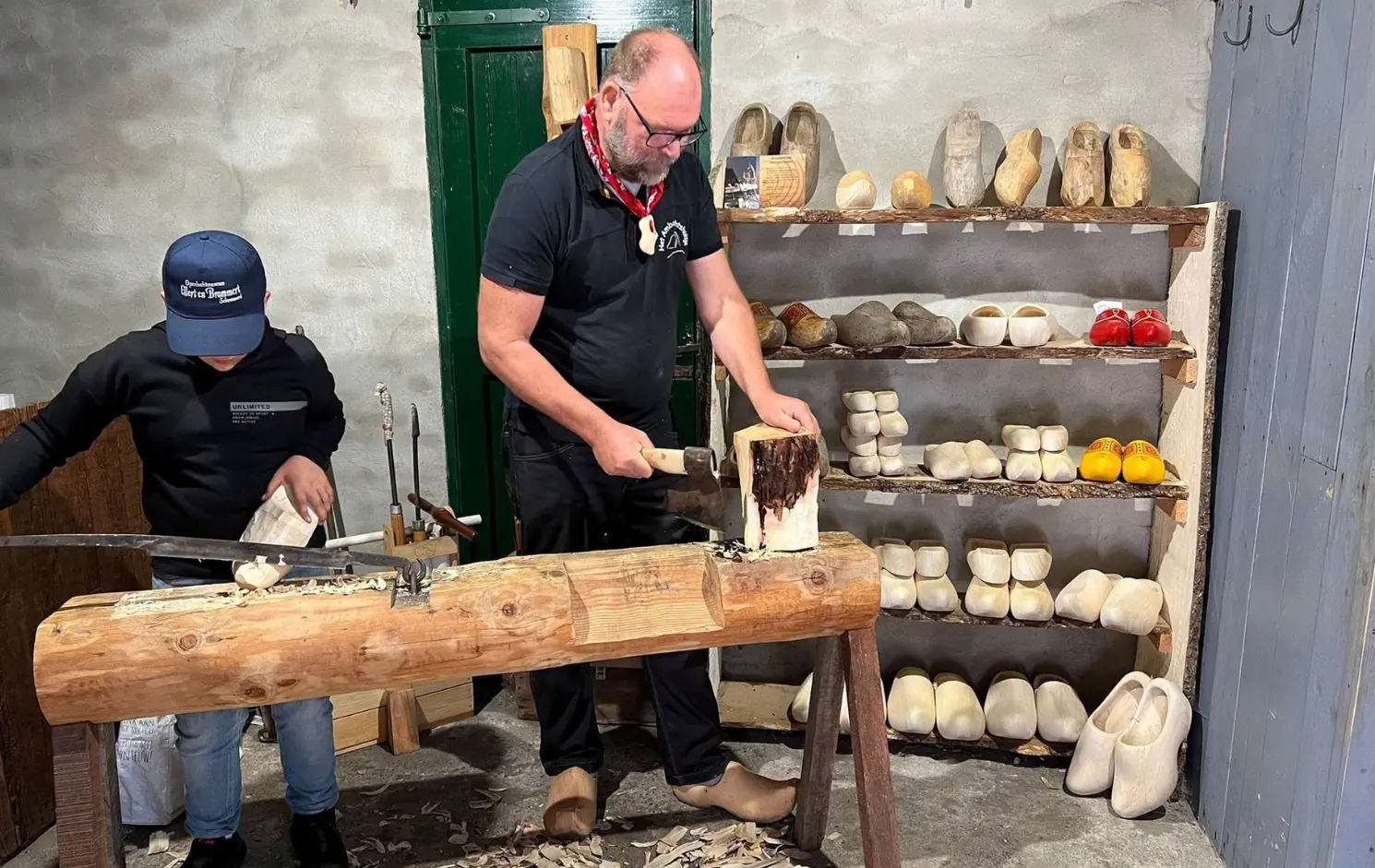 A clog maker shapes a clog on a wooden workbench, next to racks full of clogs.