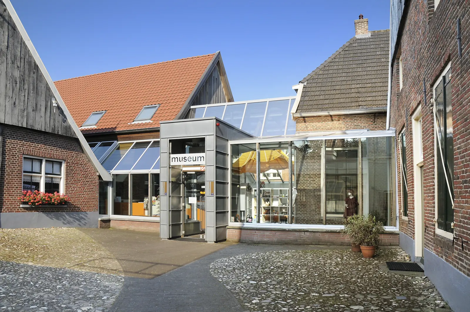 Ingang van het Nationaal Onderduikmuseum in Aalten: een glazen entree tussen oude bakstenen panden, met kinderkopjes ervoor.