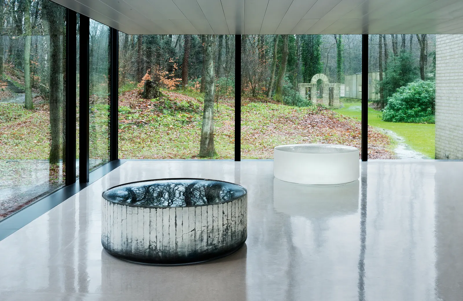 Twee ronde glazen sculpturen op de vloer van een zaal met grote ramen naar het bos.