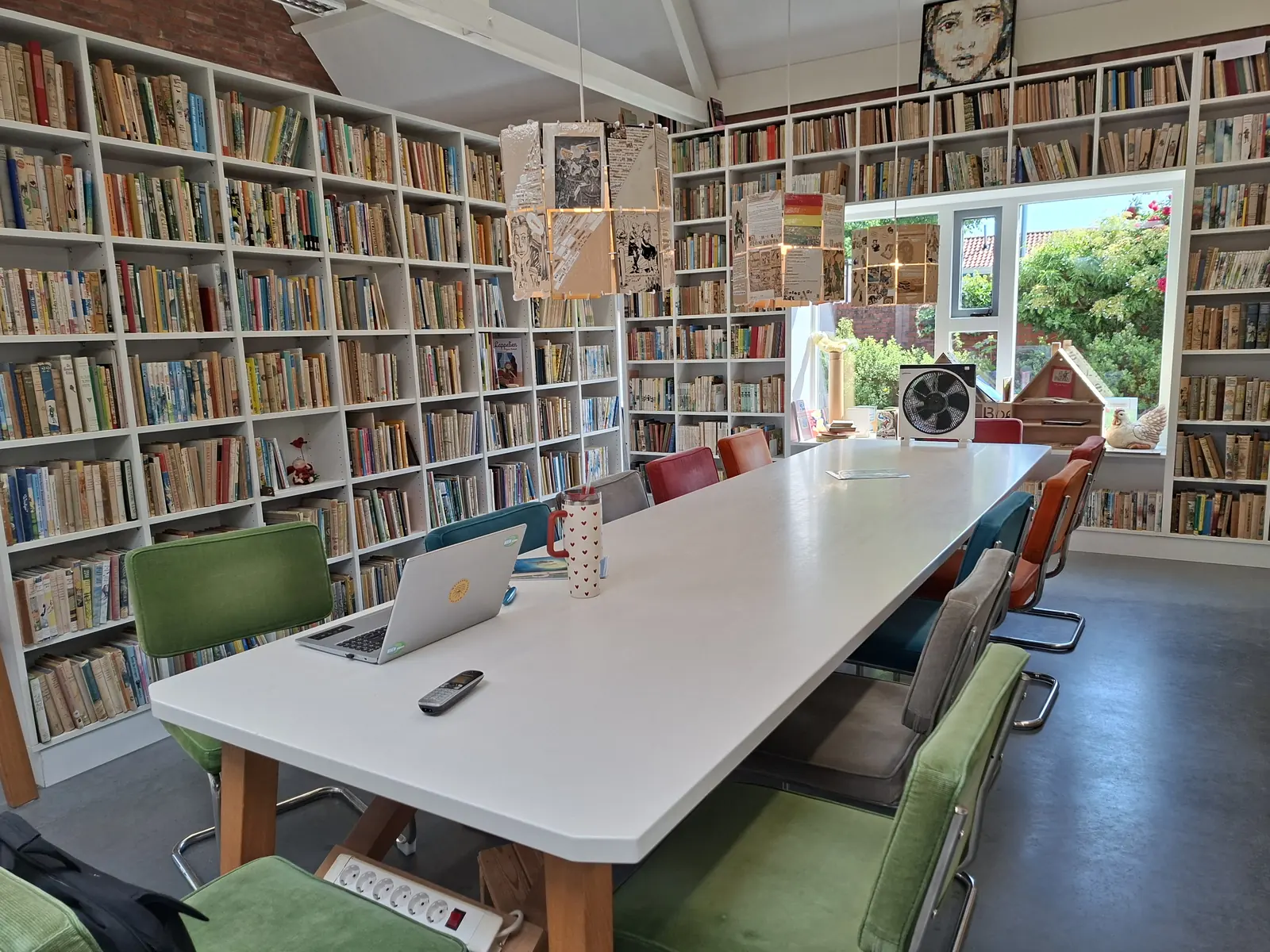 Une salle lumineuse avec une longue table blanche et des chaises vertes, entourée de hautes bibliothèques remplies de livres pour enfants.