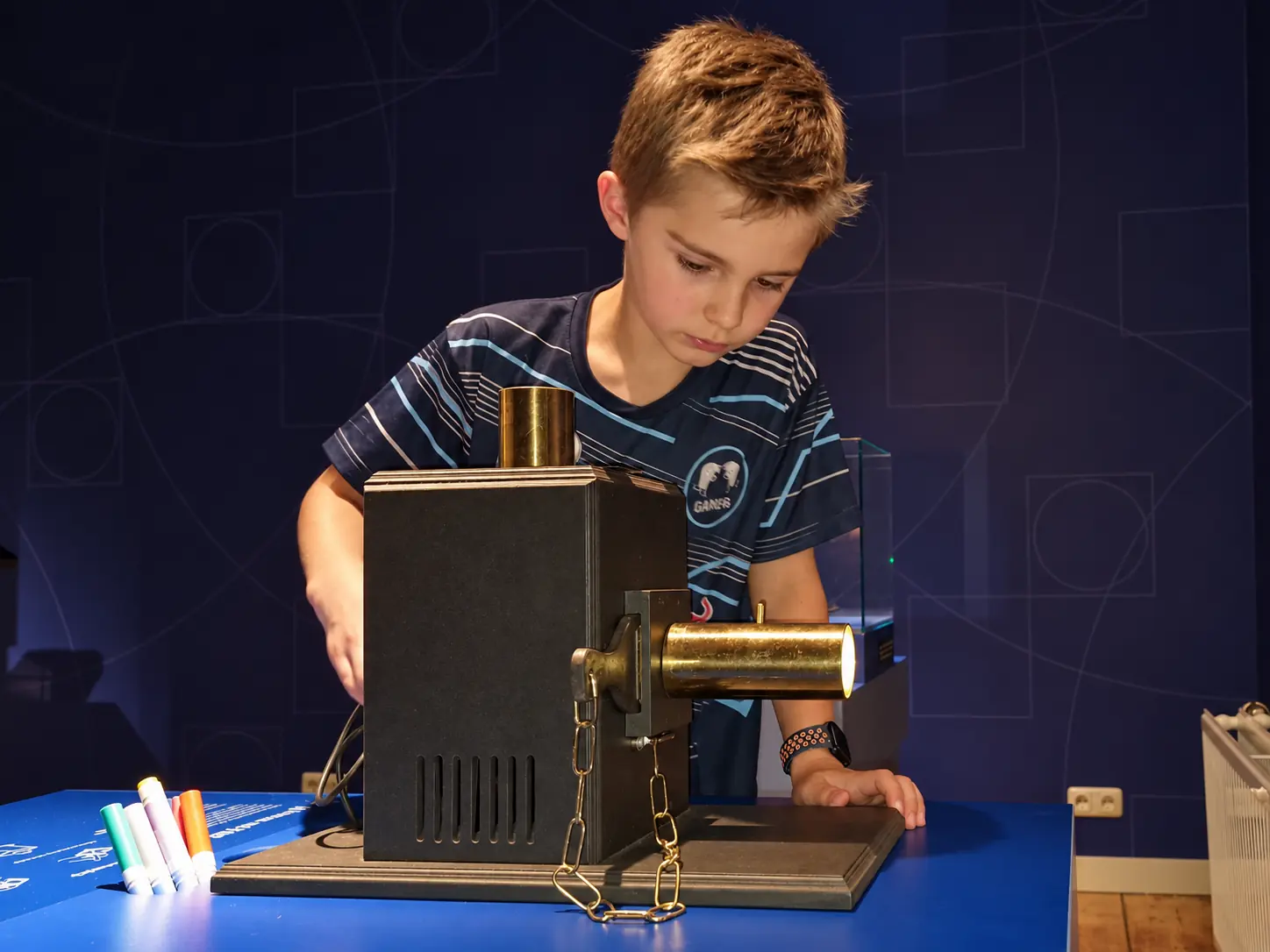 A boy operates a replica magic lantern on a table with marker pens.