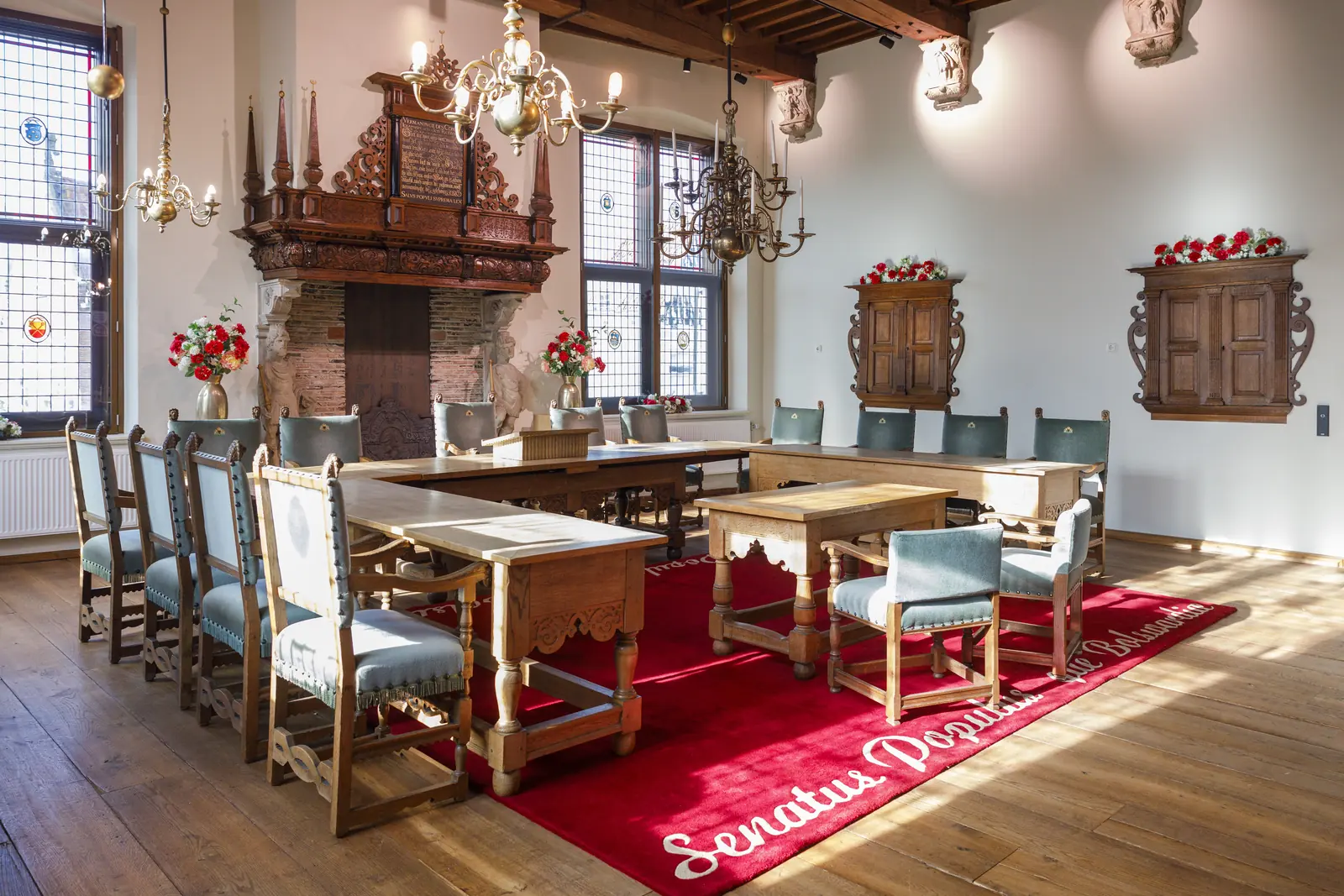 Een raadzaal met houten tafels, blauwe stoelen, kroonluchters, een rood tapijt en een rijk gesneden schouw.