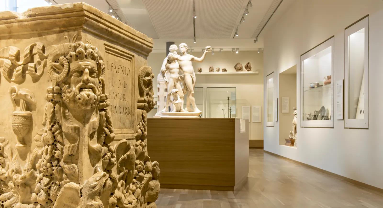 Un autel romain sculpté orné d’un visage en relief ; à l’arrière-plan, un groupe en marbre et des vitrines.