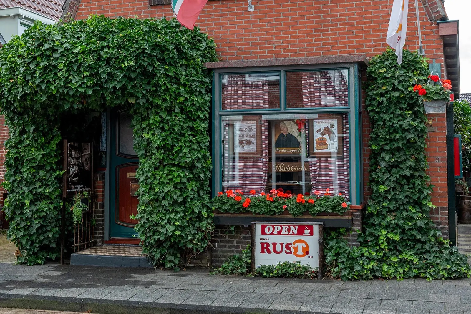 Fassade von ’t Hoeske van Opoe Iet, mit Efeu bewachsen, mit Geranien und einem Schild mit „Open“ und „Rust“.