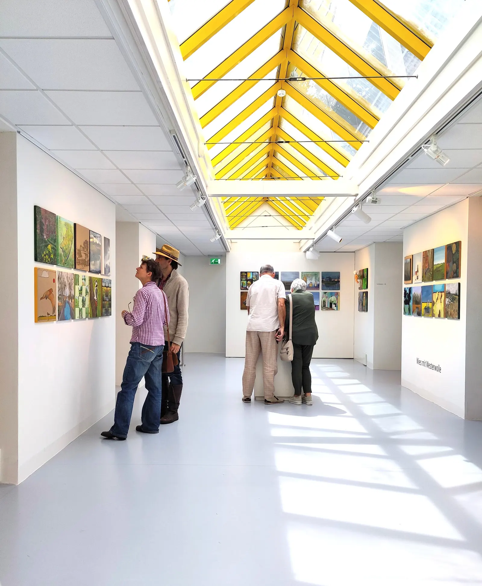 Bezoekers bekijken schilderijen in een lichte zaal met een geel glazen dak.