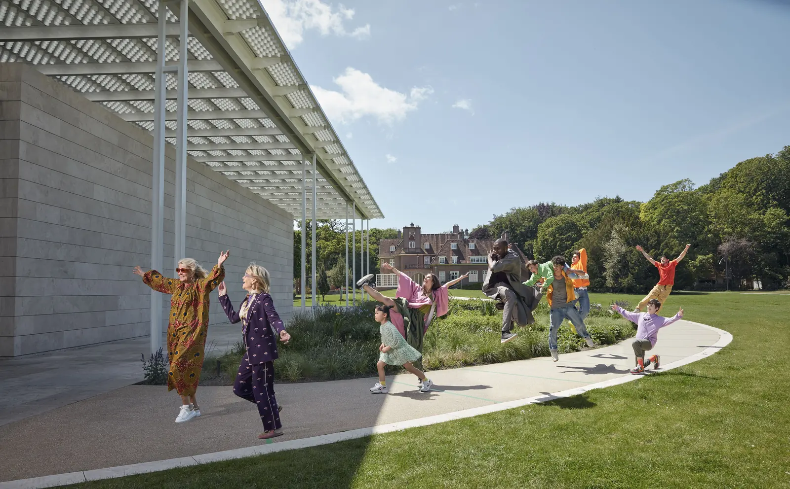 Bezoekers lopen en dansen over een gebogen pad langs het lage museumgebouw van Voorlinden, met tuin en bomen.