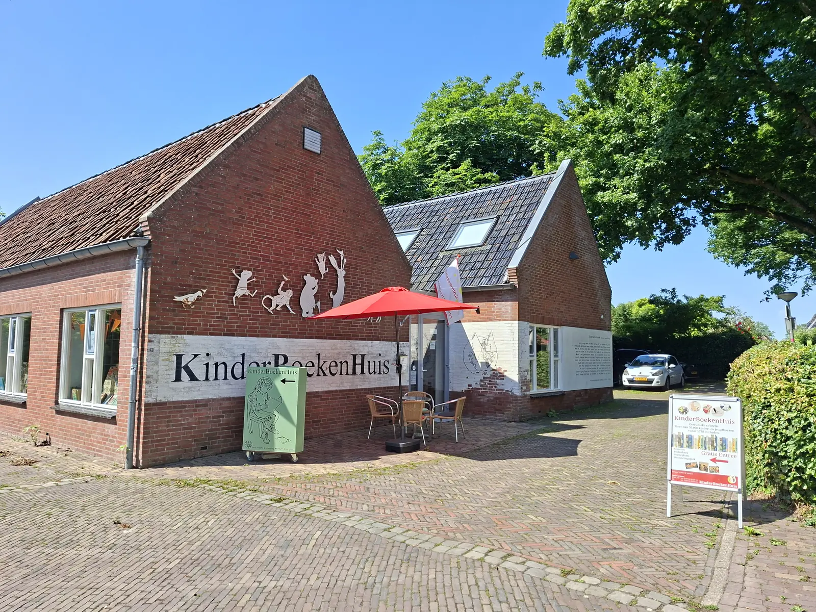 Le bâtiment bas en brique du KinderBoekenHuis, avec des silhouettes sur la façade, une terrasse sous un parasol rouge et un panneau à l'entrée.