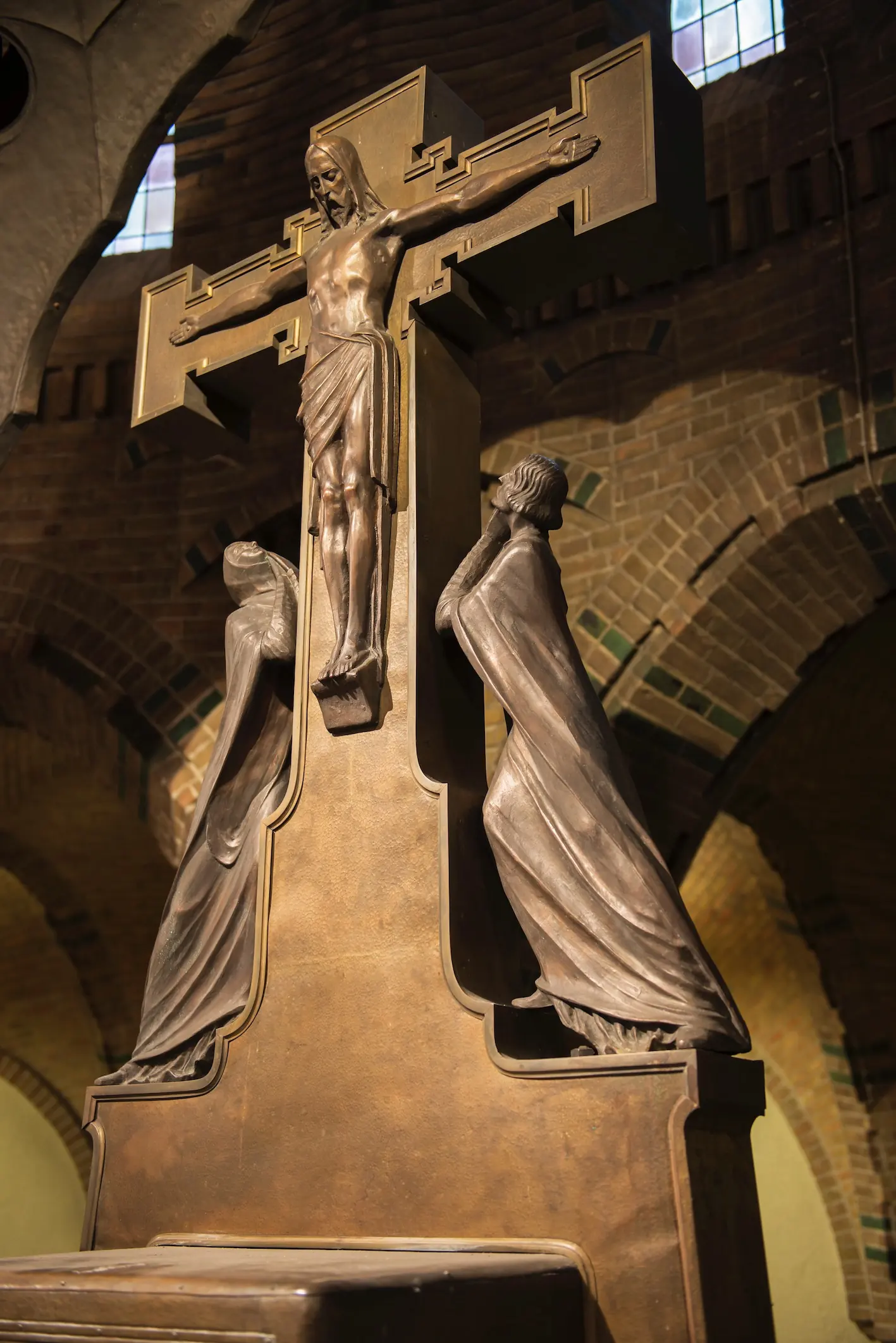 Un crucifix en bronze encadré de deux figures allongées en deuil, sur un fond d’arcs en brique.