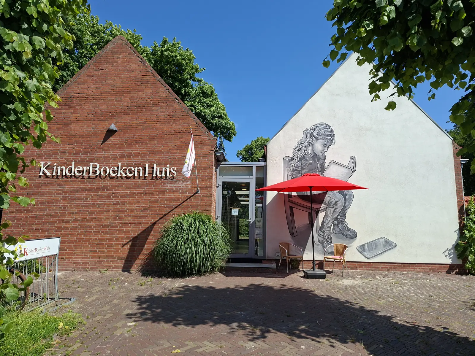 Façade en brique portant les lettres KinderBoekenHuis, à côté d'un pignon blanc orné d'une grande peinture d'une fillette qui lit, avec un parasol rouge près de l'entrée.