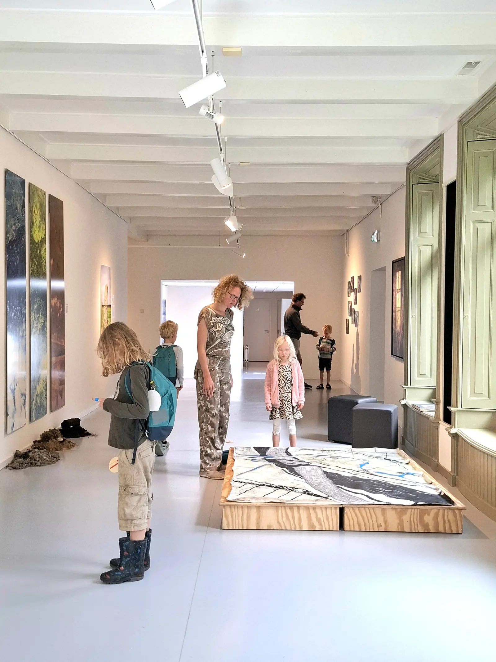 Een volwassene en kinderen bekijken een kunstwerk op een houten verhoging in een witte museumzaal van het MOW.
