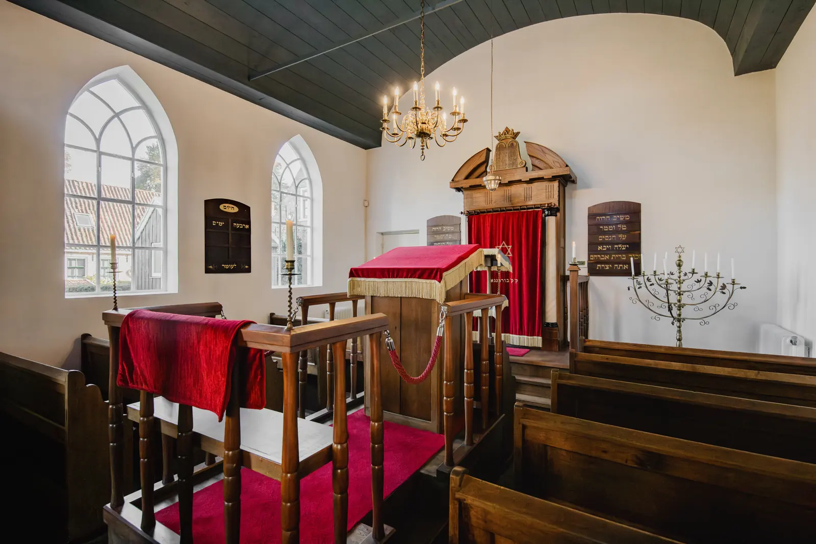 Interieur van de synagoge in Bourtange met houten banken, rode kleden, kroonluchters en een menora.
