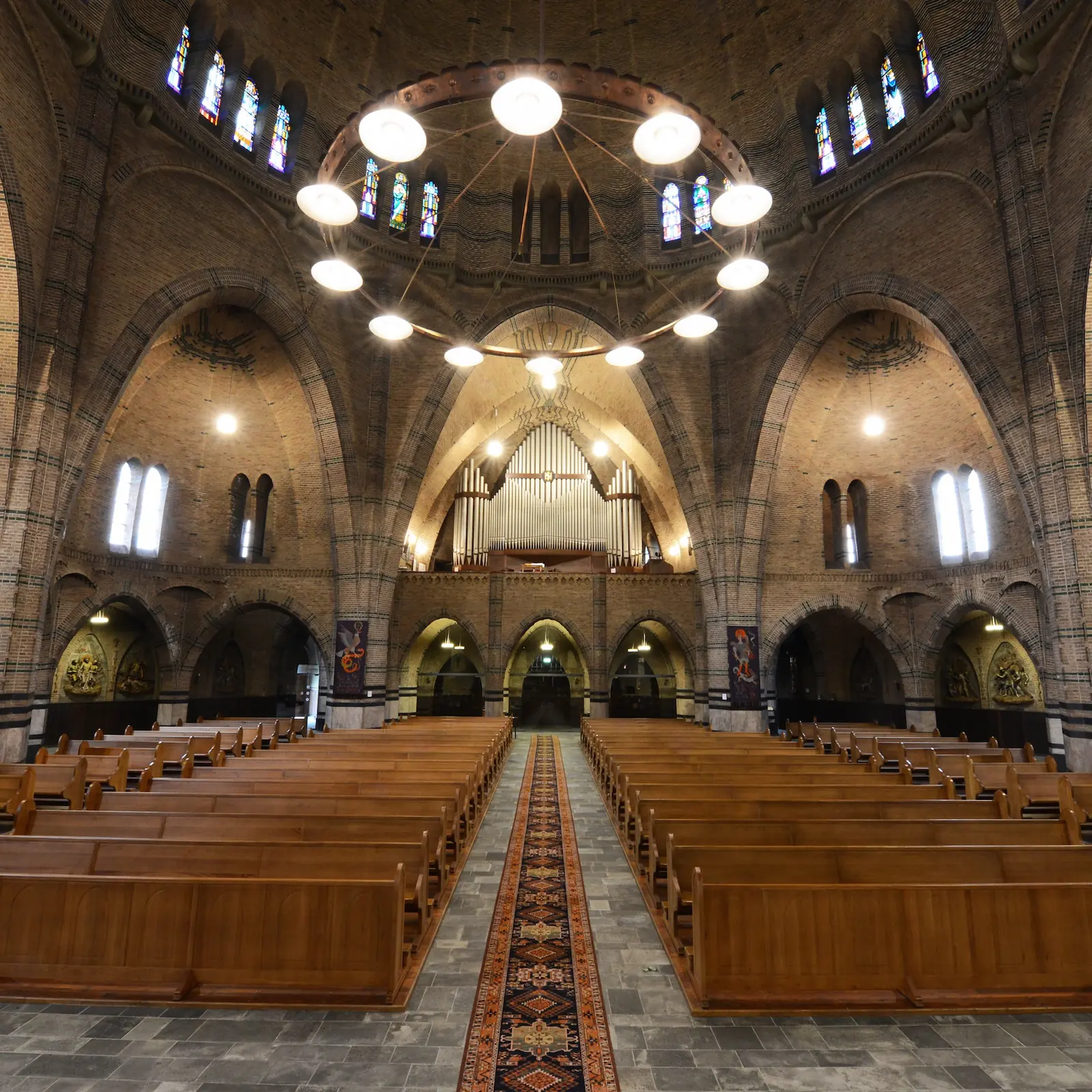 L’intérieur de l’église avec des rangées de bancs en bois et un tapis dans l’allée centrale, de hauts arcs en brique, une couronne de lampes sous la coupole et l’orgue au fond.