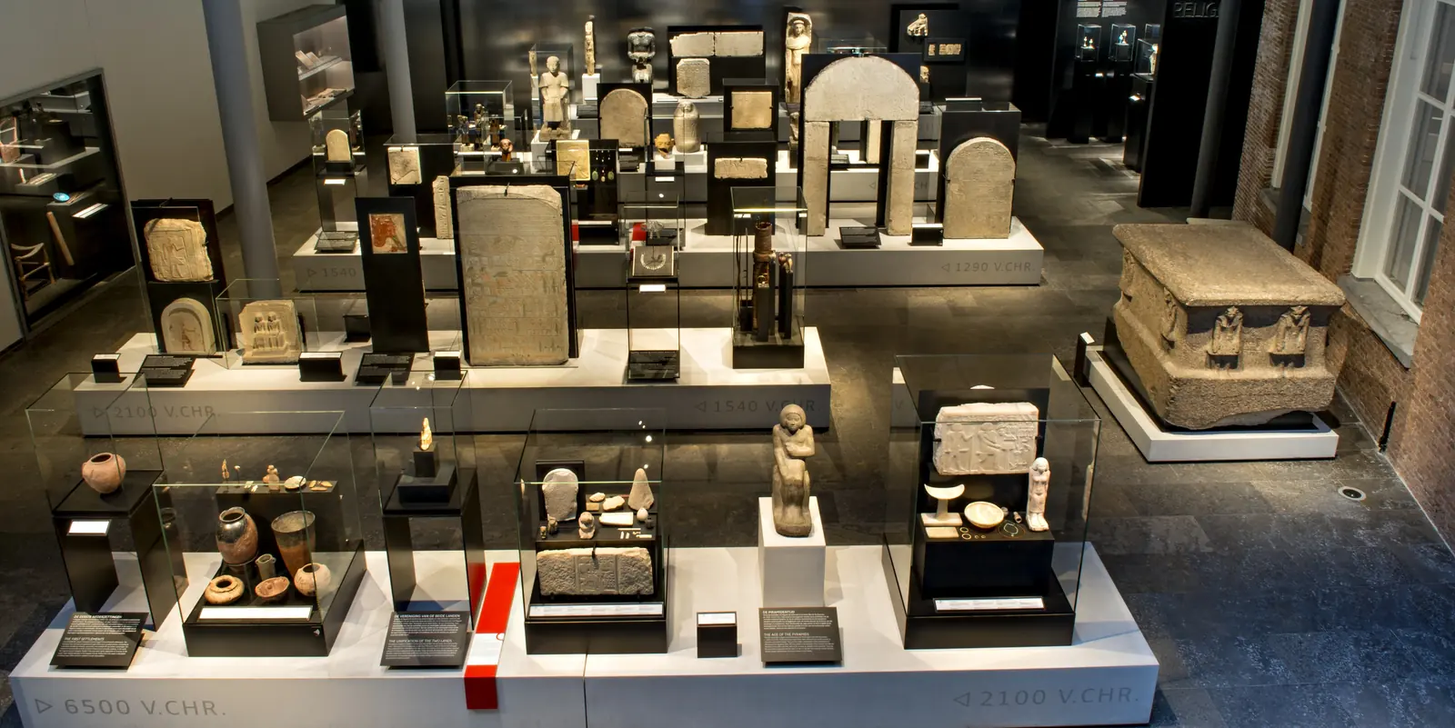 Une salle avec des stèles, des statuettes et un sarcophage sur des socles bas éclairés.