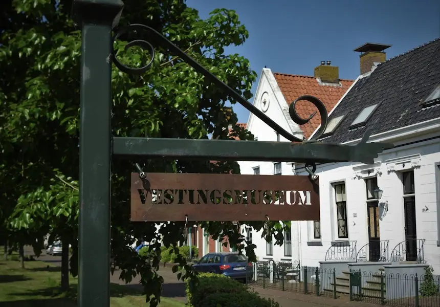 Een uithangbord met het opschrift Vestingsmuseum aan een smeedijzeren arm, met daarachter witte huizen en bomen.