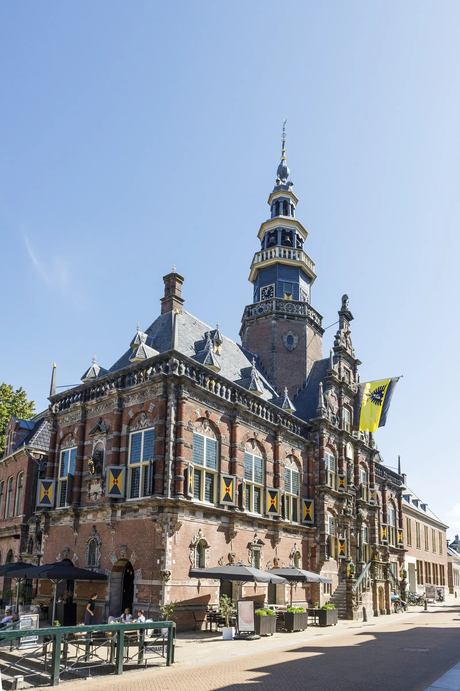 Het historische stadhuis van Bolsward met een rijk versierde gevel, een hoge toren en een vlag, op een zonnige dag.