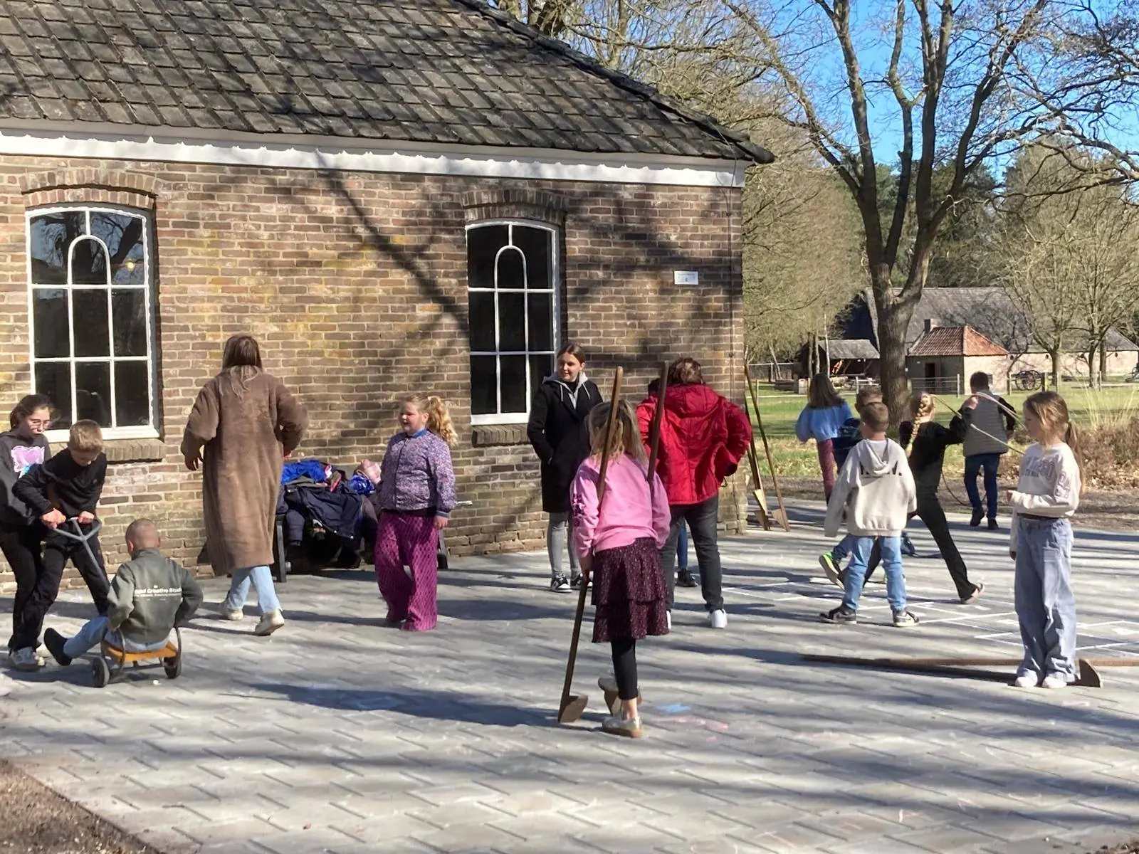 Children play with old-fashioned toys in a paved schoolyard in front of a brick school building.