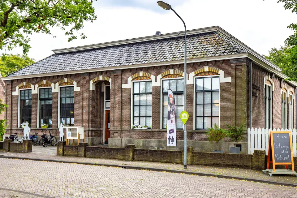 De bakstenen gevel van de oude Scheepstraschool met hoge ramen onder lichte bogen, een schilddak en een klinkerstraat ervoor; links staan fietsen, rechts een stoepbord.