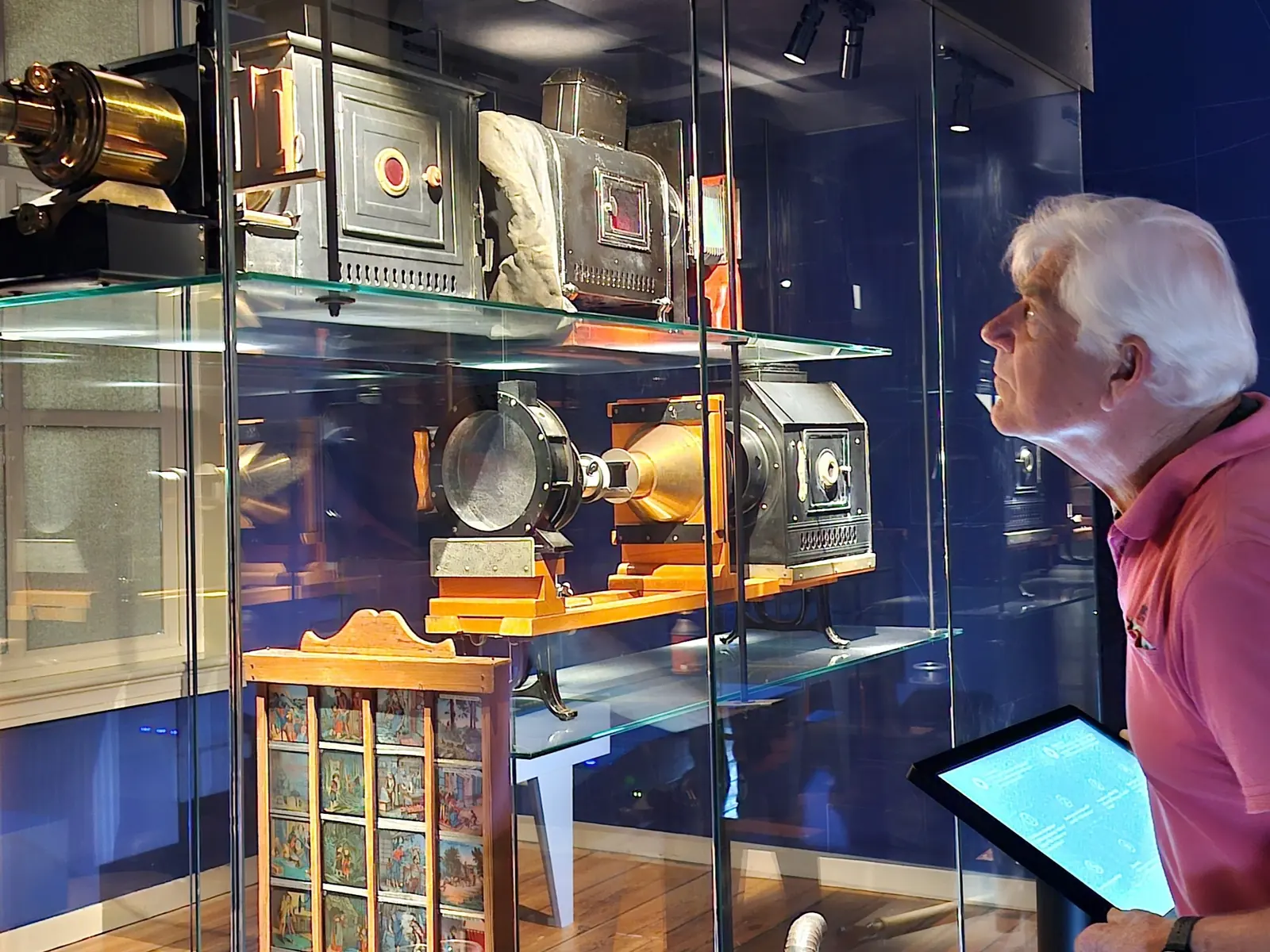 A visitor holding a tablet looks at a display case of old magic lanterns and painted glass slides.