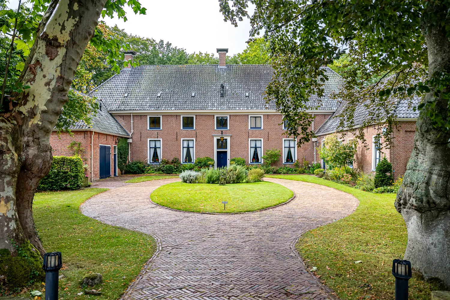 Façade de Havezate Mensinge avec un jardin rond et une allée pavée entre de grands arbres.