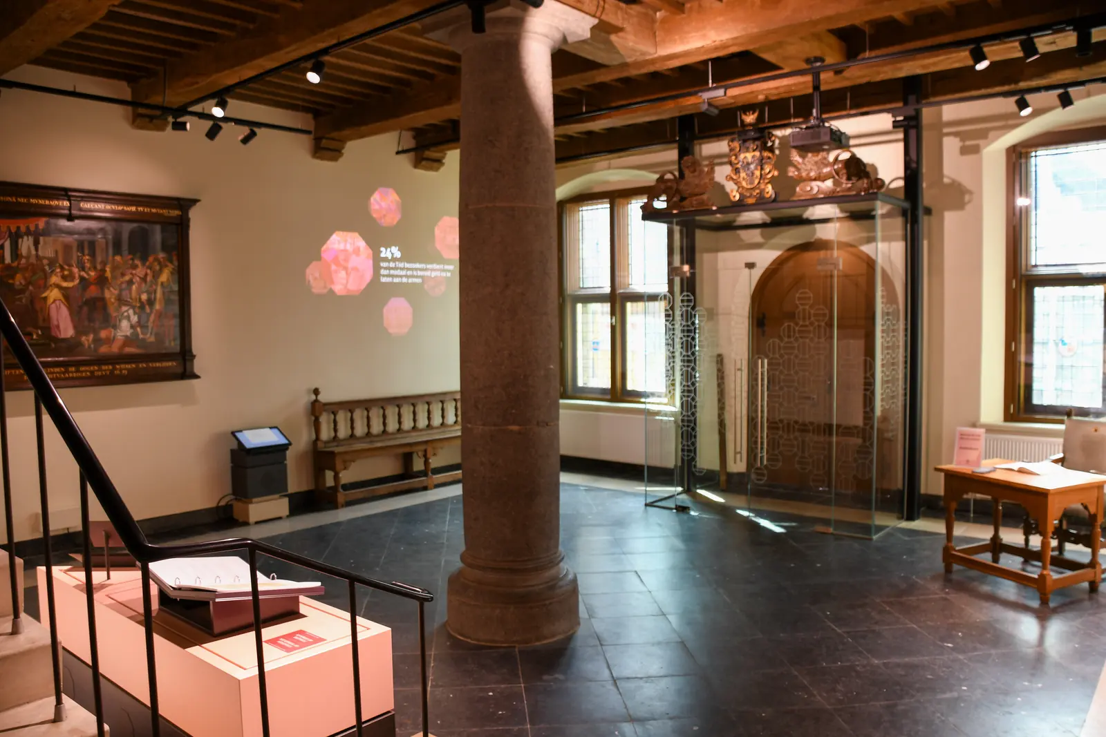 Een tentoonstellingsruimte met een stenen zuil, een schilderij, een vitrine en een houten balkenplafond.