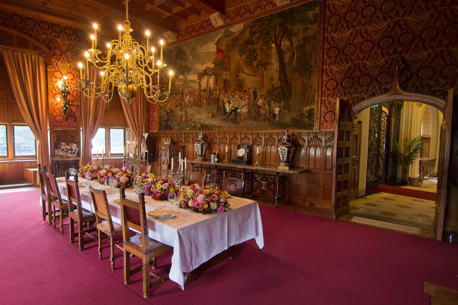 Een gedekte tafel in een eetzaal met houten lambrisering, een kroonluchter en grote wandschilderingen.