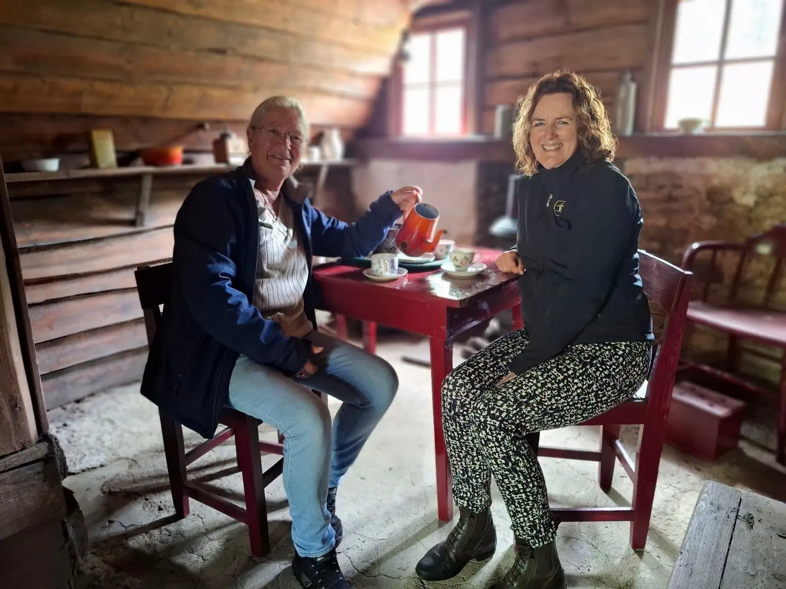 Two women drinking coffee at a small red table in a simple wooden hut.
