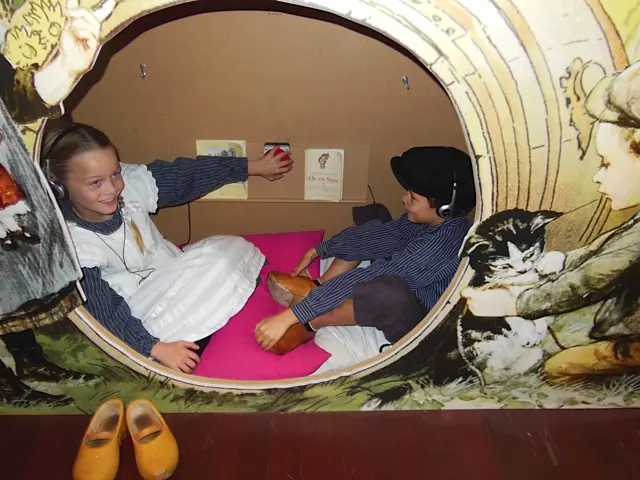 Een meisje met een wit schort en een jongen met een pet, verkleed als Ot en Sien, zitten met koptelefoons op in een ronde nis die als ton is beschilderd; ervoor staan gele klompjes.