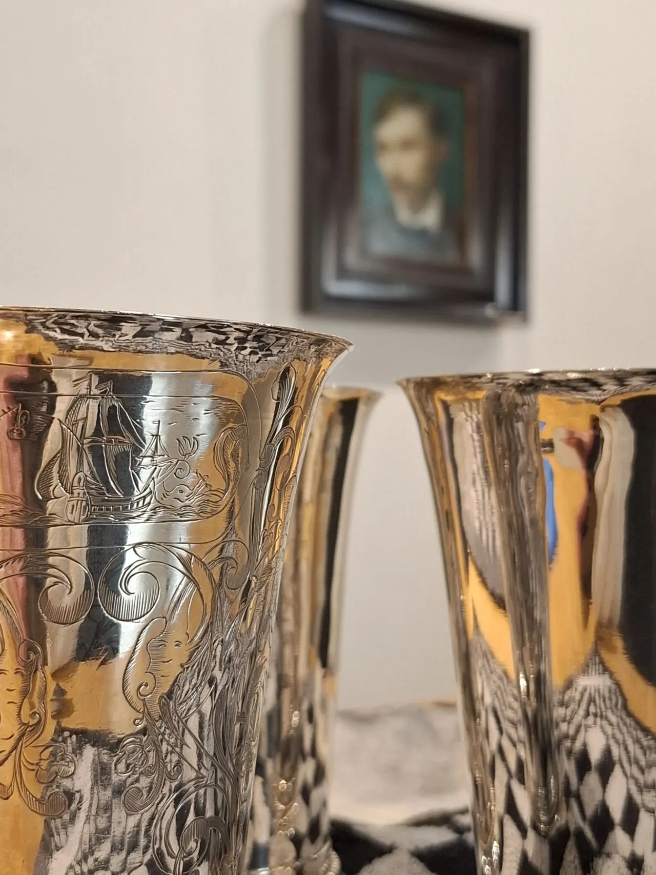 Two decorated silver cups with a painting in the background.