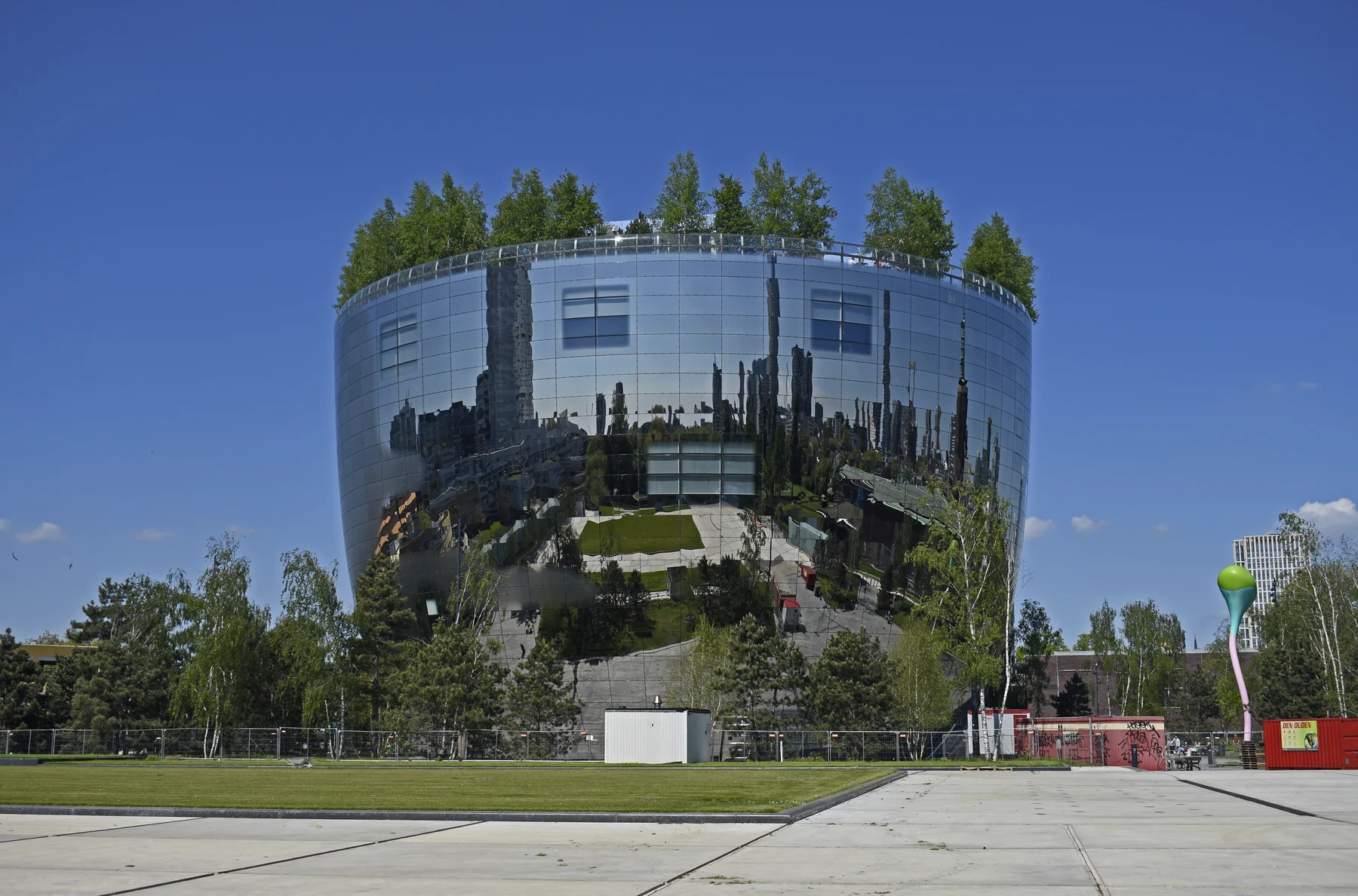 Round mirrored depot building with trees on its roof and a paved square in front.