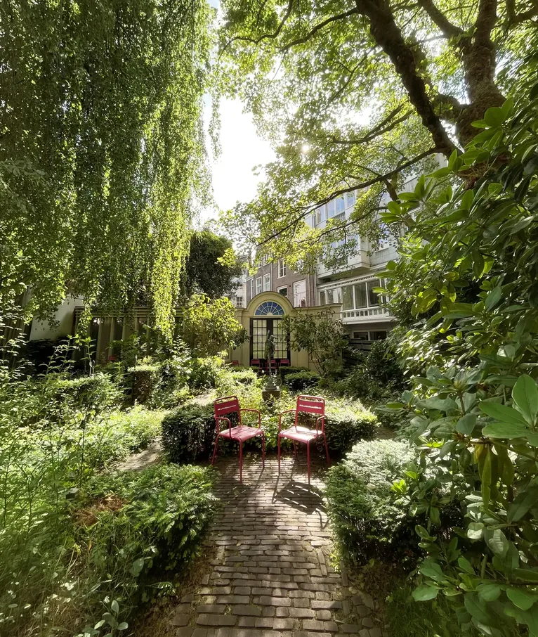 Green canal garden with a brick path and two red chairs.