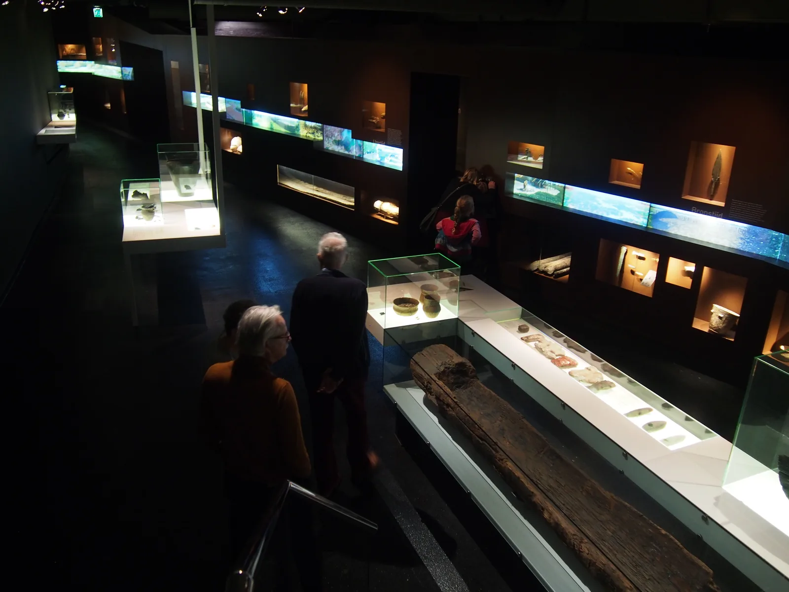 Dark museum gallery with visitors and the long wooden Pesse canoe in an illuminated case.