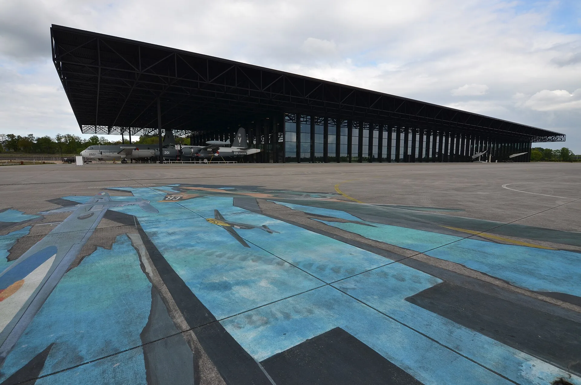 The wide museum building with aircraft under its canopy and painted paving in the foreground.
