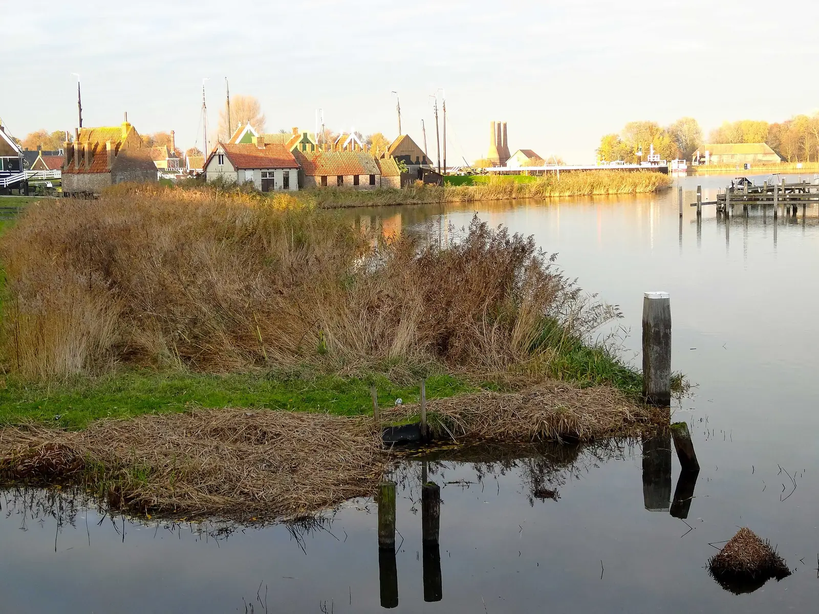 Tiled-roof houses beside a wide stretch of water, with reeds and boat masts behind them.